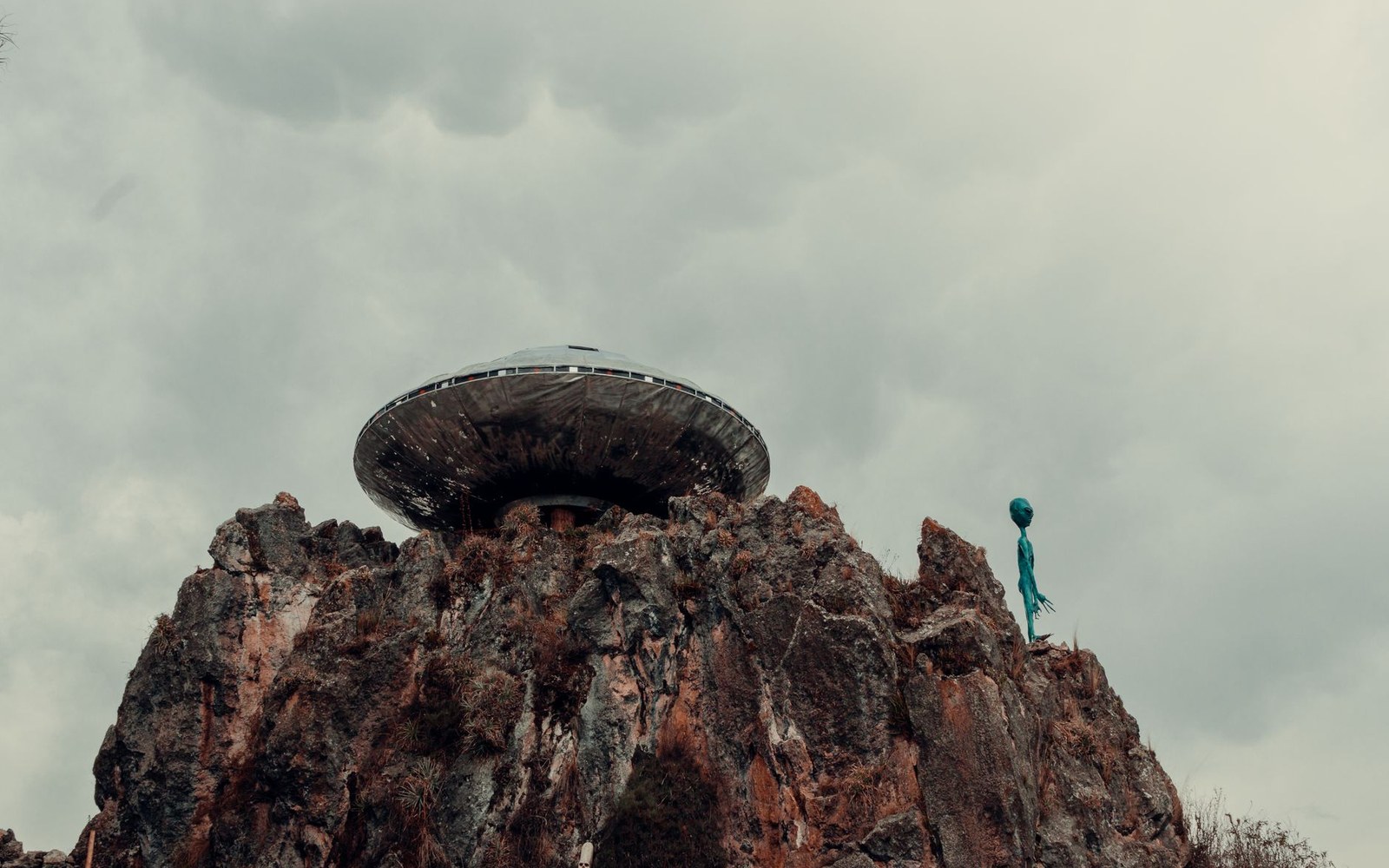 A saucer-shaped structure and alien figure statue perched atop a rocky peak under grey skies