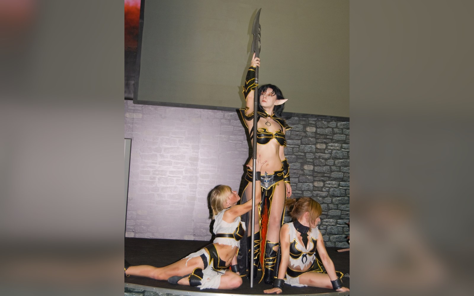 Cosplayers in elf-warrior costumes pose on stage with one holding a large blade staff