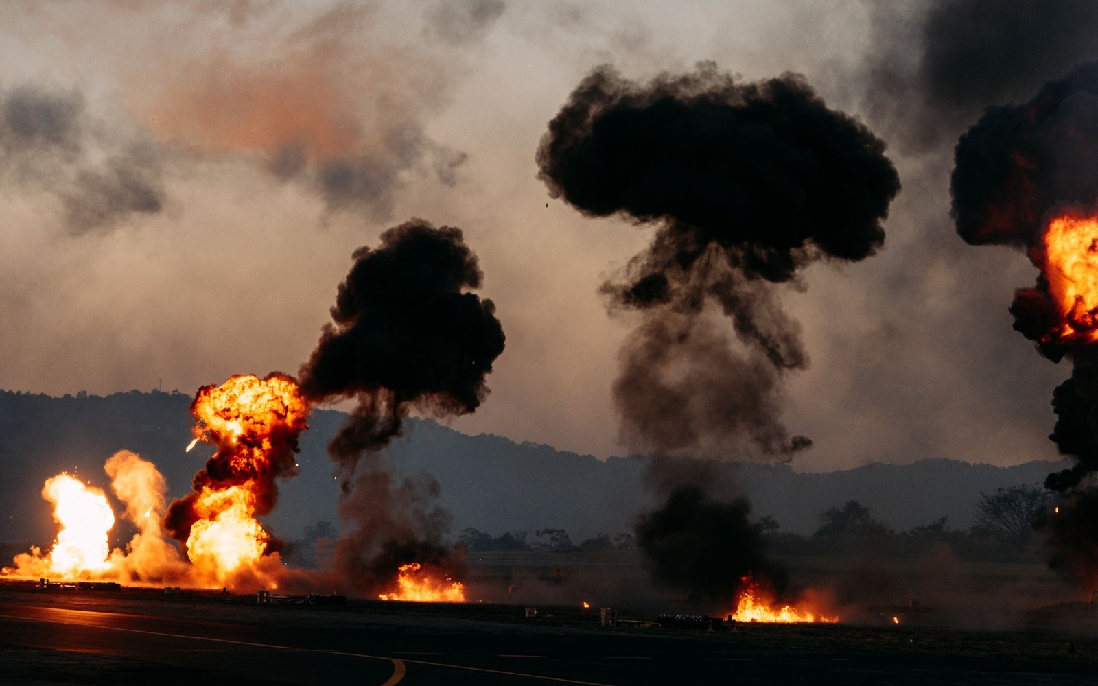 Multiple fiery explosions send dark mushroom clouds of smoke into the sky over a runway