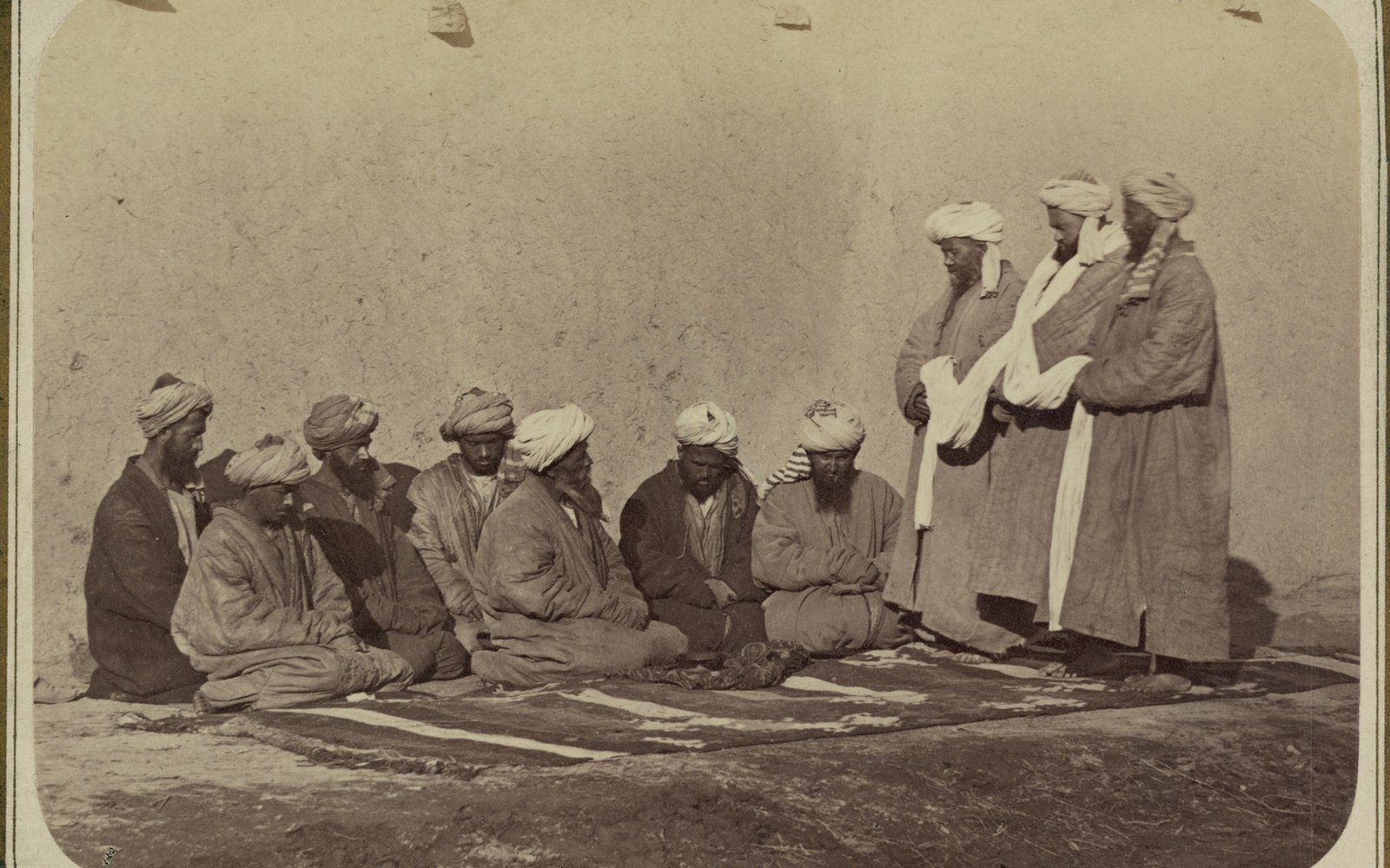 Seated men in turbans and robes on a carpet face three standing men against a wall
