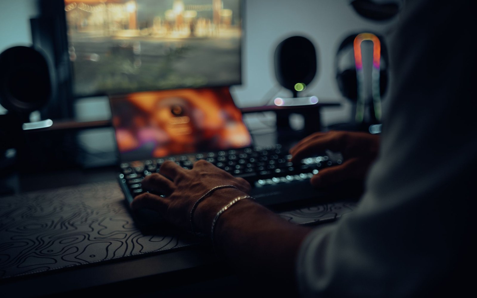 A desktop monitor in a darkened room showing the World of Warcraft retail UI during a raid encounter, the screen the only light source, an adult player's hands visible on the keyboard below
