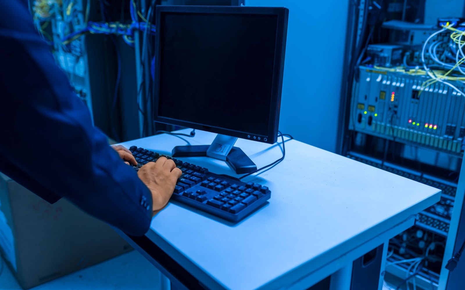 A row of illuminated server racks in a data-centre corridor, an adult technician in the background